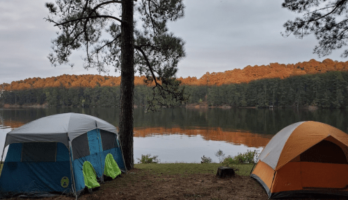 Two tents by a calm lake, surrounded by trees, with a colorful forest reflecting on the water's surface.