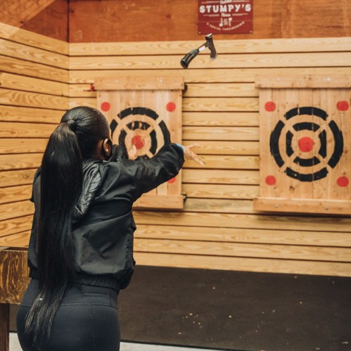 A person throws an axe at a target in a wooden axe-throwing venue with a sign reading "Stumpy's."