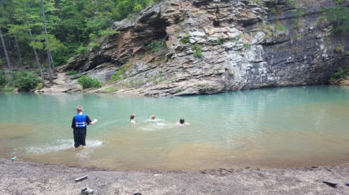 A child in a life jacket stands by a rocky shore while three kids swim in a clear, greenish-blue water.