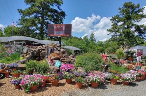 A vibrant garden center with colorful flowers, a waterfall feature, and a digital sign under a blue sky with fluffy clouds.