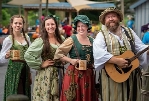 Four people in medieval costumes pose together, holding mugs and a guitar, in a festive outdoor setting.