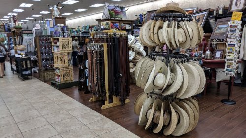 A store interior featuring racks of hats, belts, and various souvenirs, with shoppers browsing in the background.