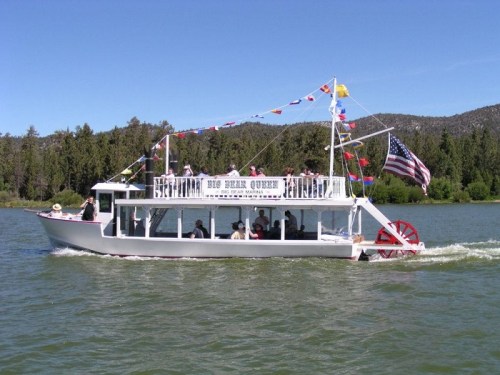 A paddleboat named "Big Bear Queen" cruises on a lake, decorated with flags and an American flag, surrounded by trees.
