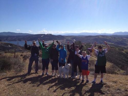 A group of eight people and a dog stand on a hillside, raising their arms in celebration against a scenic backdrop.