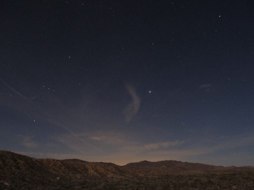 A night sky filled with stars over a desert landscape, with mountains silhouetted against the horizon.