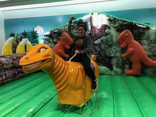A person joyfully rides a bright orange dinosaur on a green inflatable surface, surrounded by inflatable dinosaur decorations.