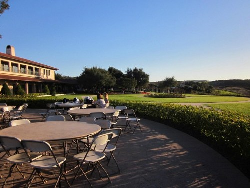 A sunny outdoor setting with empty tables, a green lawn, and a building in the background.