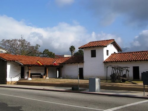 Historic building with white walls and red-tiled roofs, set against a cloudy sky and surrounded by greenery.