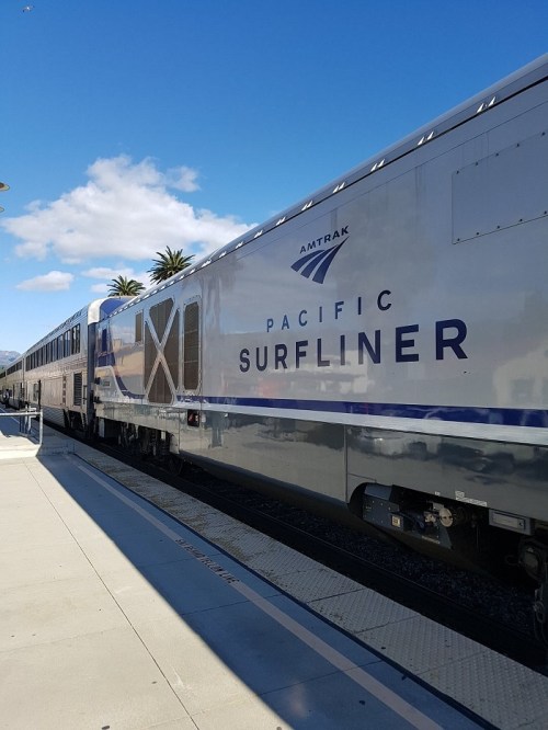 Amtrak Pacific Surfliner train at a station, with palm trees and blue sky in the background.