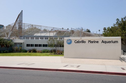 Sign for Cabrillo Marine Aquarium with a modern building and greenery in the background on a clear day.