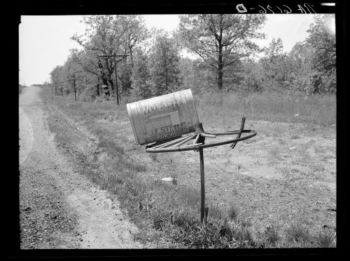A weathered mailbox on a metal post beside a dirt road, surrounded by trees and open fields.