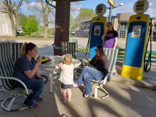 Two women and a child sit at a table outside a vintage gas station, enjoying drinks in the sunlight.