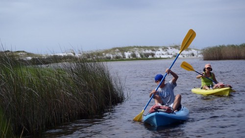 Two people kayaking on a calm waterway surrounded by tall grass and sandy hills under a cloudy sky.