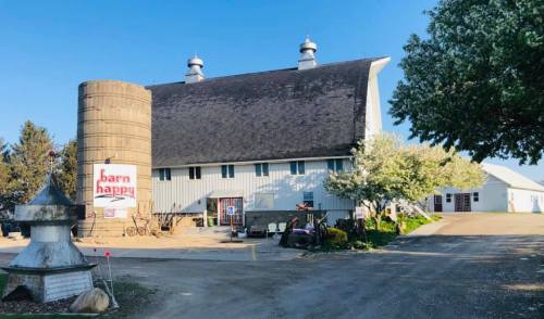 A large barn with a silo, surrounded by trees and a clear blue sky, featuring a sign that says "barn happy."