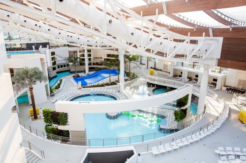 A spacious, modern resort pool area featuring multiple pools, lounge chairs, and tropical plants under a bright, open roof.