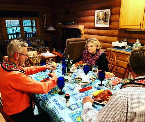 Three people dining at a cozy log cabin table, enjoying a meal with drinks and a festive table setting.