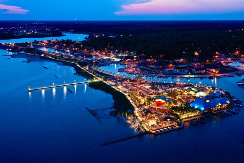Aerial view of a vibrant waterfront area at dusk, with lights reflecting on the water and boats docked nearby.