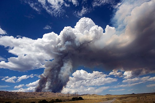 A dramatic sky filled with dark smoke billowing from a wildfire, contrasting against fluffy white clouds and blue sky.