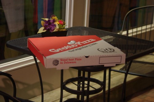 A Grotto Pizza box sits on a table outside, with a small flower pot nearby.