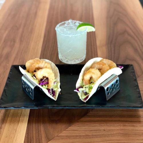 Two shrimp tacos in metal holders on a black plate, accompanied by a margarita with a lime wedge.