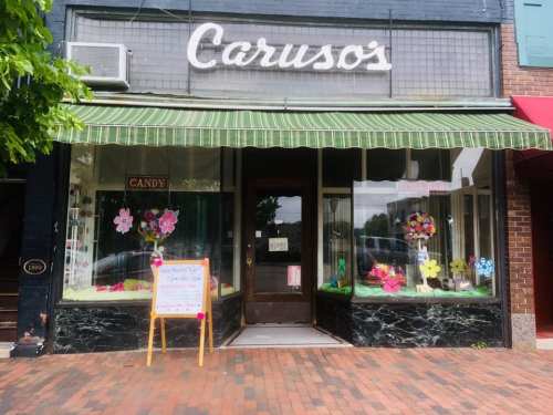 A storefront with a green awning, featuring colorful flower decorations and a sign that reads "Open" at Caruso's Candy.