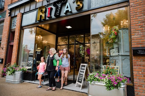 A group of four women and girls exit a shop called Rita's, surrounded by colorful flower planters.