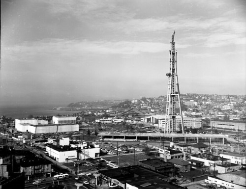 A black and white aerial view of a cityscape featuring a tall tower, buildings, and a coastal background.