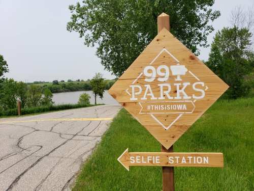 Wooden sign for "99 Parks" with a selfie station arrow, near a river and green landscape in Iowa.