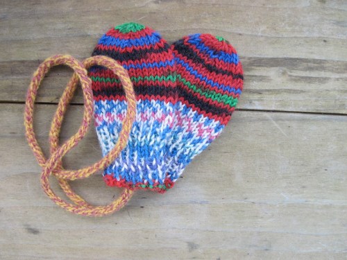 Colorful knitted mittens in the shape of hearts, with a braided cord, resting on a wooden surface.