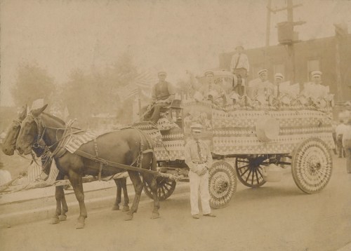 A vintage parade scene featuring a decorated horse-drawn wagon with people in uniforms and American flags.