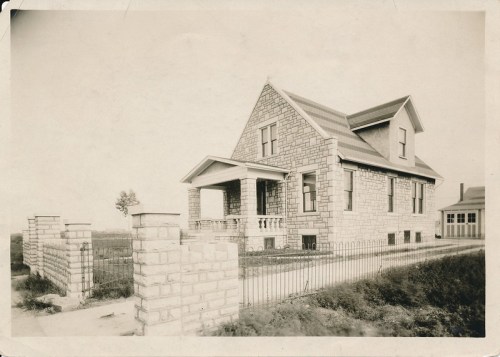 Historic stone house with a gabled roof, front porch, and iron fence, set in a grassy area. Black and white photo.
