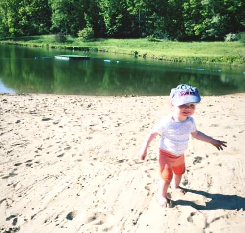 A toddler in a hat and shorts walks on a sandy beach near a calm lake surrounded by greenery.