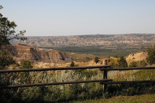 A scenic view of rolling hills and valleys under a clear sky, framed by a wooden fence and lush greenery.