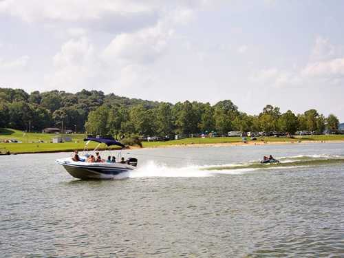 A boat with passengers speeds across a lake, while a person on a jet ski follows closely behind. Lush greenery in the background.