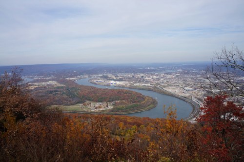 A scenic view of a river winding through a city, surrounded by autumn foliage and a cloudy sky.