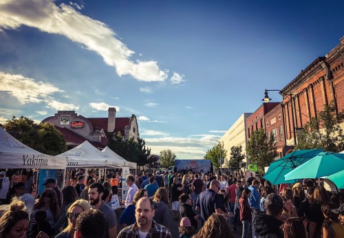 A bustling street fair with crowds, tents, and blue umbrellas under a clear sky.