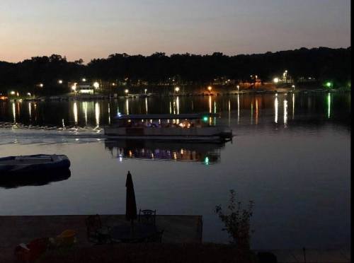 A boat with lights glides across a calm lake at dusk, reflecting colorful lights from the shore.