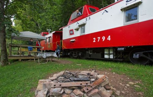 A red train caboose parked in a wooded area, with a picnic table and fire pit nearby. People are gathered outside.