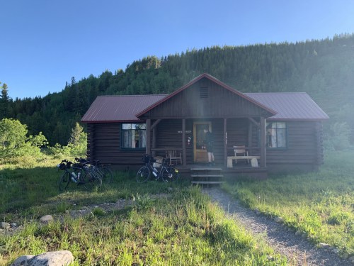 A rustic log cabin with a red roof, surrounded by greenery and bicycles parked in front on a sunny day.