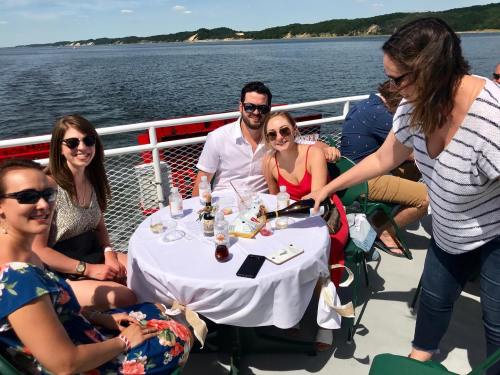 A group of five friends enjoying drinks on a boat, with scenic water and hills in the background.