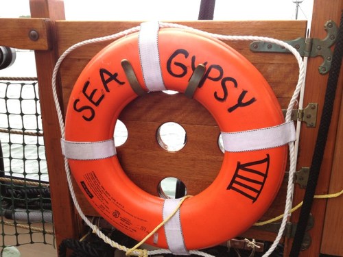 An orange life ring labeled "SEA GYPSY" hangs on a wooden ship railing near the water.