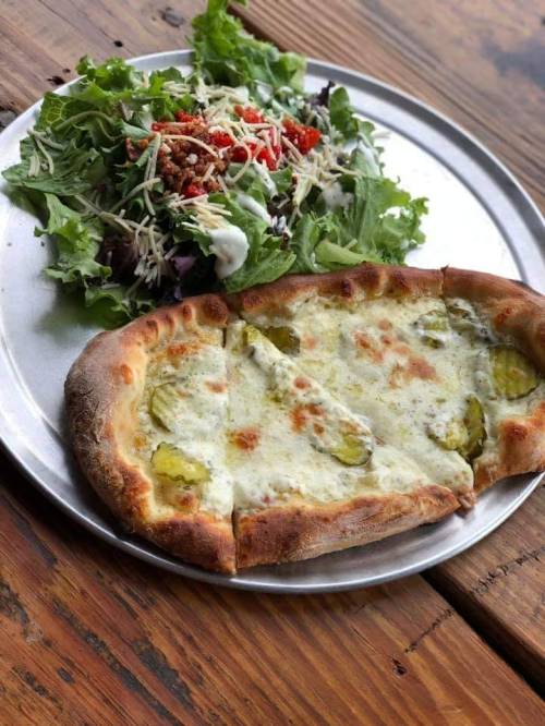A plate with a slice of cheesy pizza topped with pickles and a side salad with greens, cheese, and dressing.