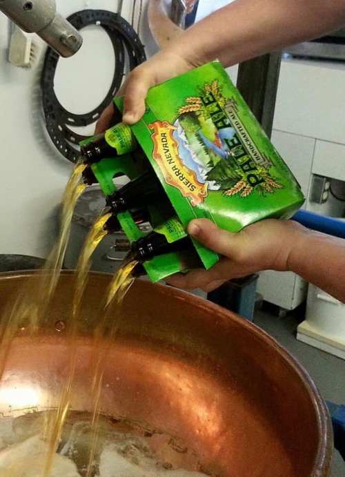 A person pours multiple bottles of beer from a six-pack into a large copper brewing kettle.
