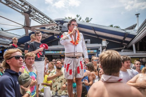 A lively outdoor party scene with a man in a white shirt and lei speaking into a microphone, surrounded by a cheering crowd.