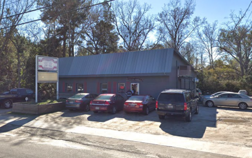 A gray building with a metal roof, featuring several parked cars in front and trees in the background.