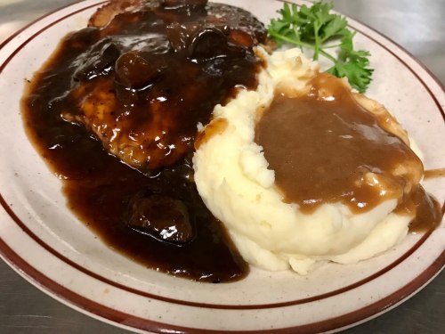A plate of meat with brown gravy and mushrooms, served alongside creamy mashed potatoes and a sprig of parsley.