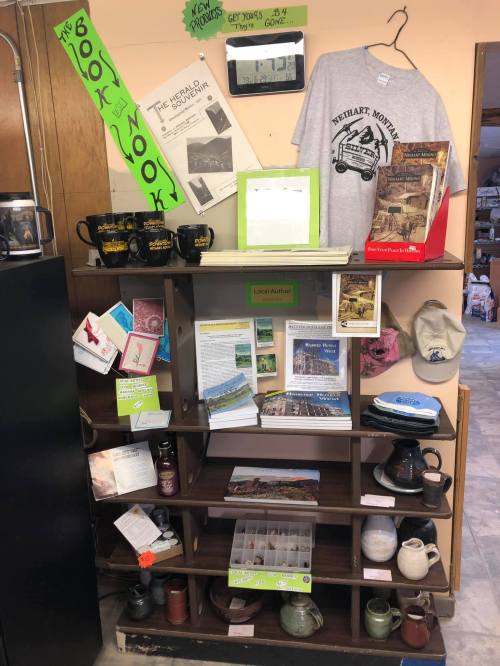 A wooden shelf displaying books, mugs, a t-shirt, and local author materials in a cozy shop setting.