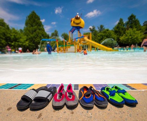 Colorful water shoes lined up on the edge of a pool, with a water slide and people enjoying the water in the background.