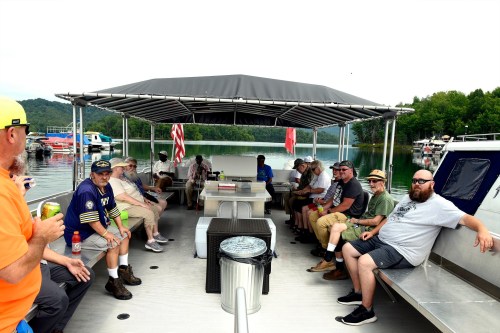A group of people sitting on a covered boat, enjoying a day on the water with trees and hills in the background.