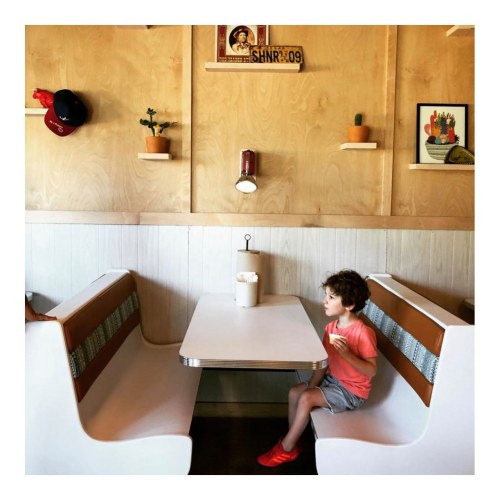 A child sits alone at a small table in a cozy diner with wooden walls and decorative shelves.